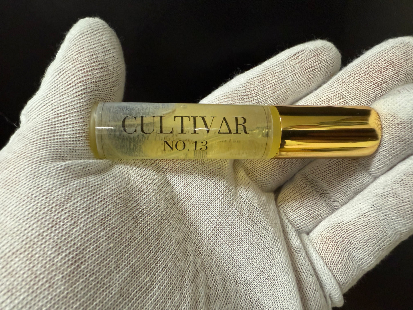 Gold tube labeled 'Cultivar No. 13' held in a white gloved hand against a black background