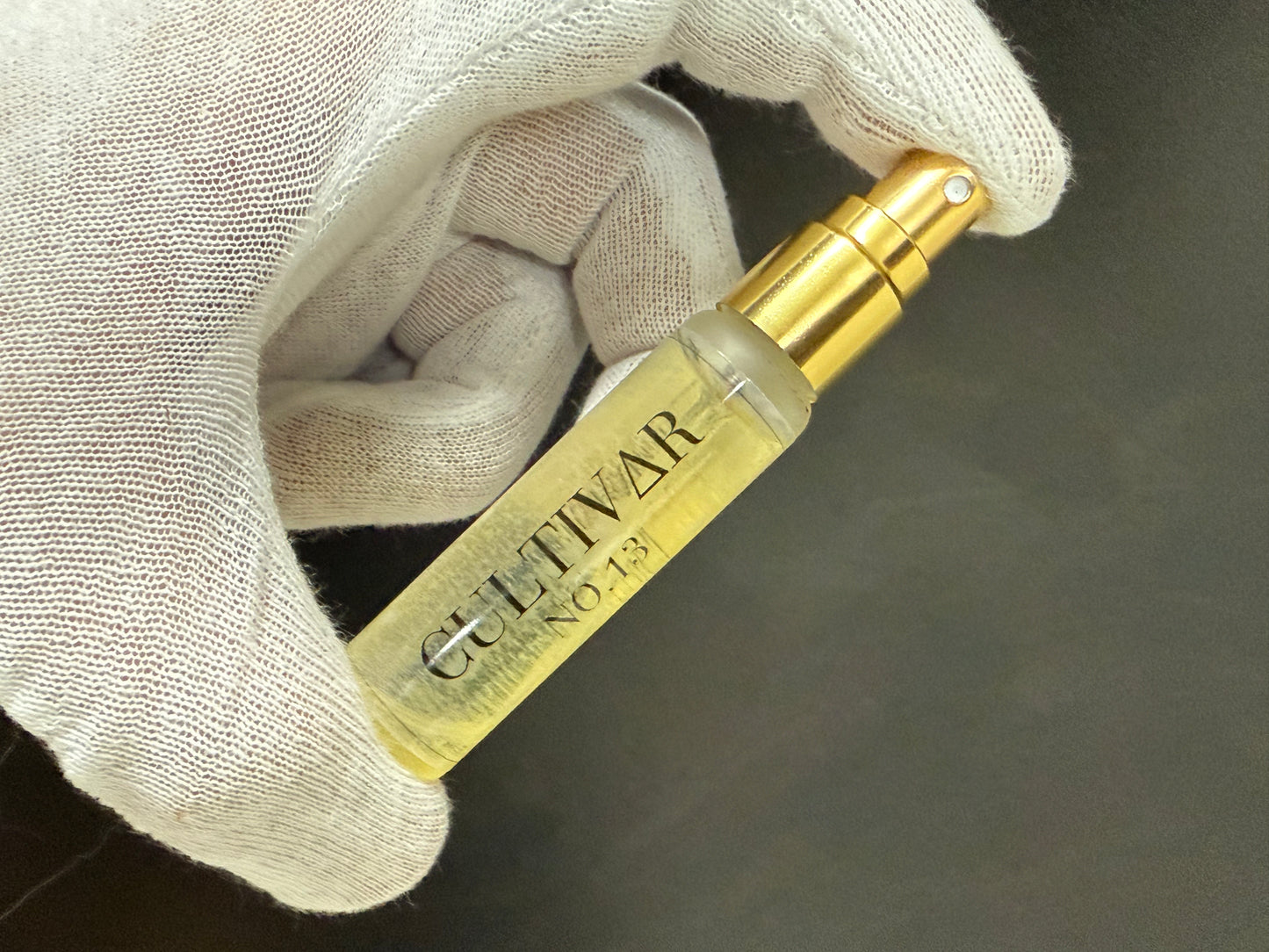 Gold bottle labeled 'Cultivar' held by a gloved hand against a dark background side view